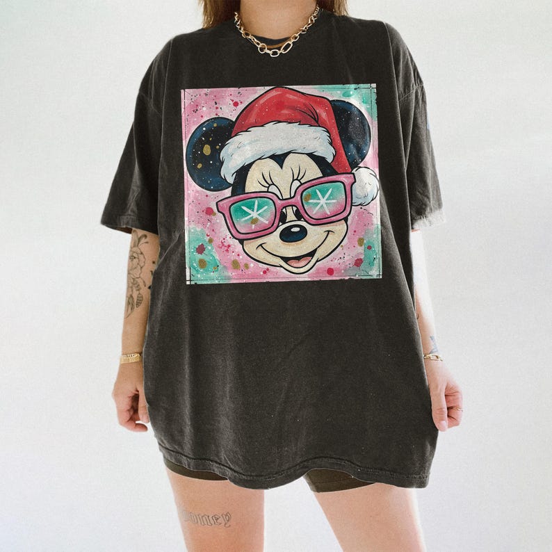 May include: Dark gray t-shirt featuring a colorful graphic of Mickey Mouse wearing a Santa hat and pink sunglasses with snowflake lenses. The graphic is set against a pink and teal speckled background. The shirt has short sleeves and a relaxed fit.