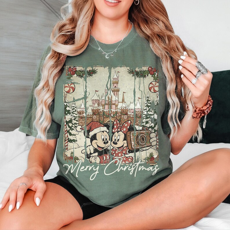 May include: Green t-shirt featuring Mickey and Minnie Mouse in front of a castle, holding a camera, with the text "Merry Christmas." The design includes Christmas trees, candy canes, and festive accents.