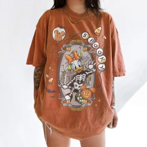 May include: An orange t-shirt featuring a Halloween-themed graphic. The design includes a skeleton Daisy Duck, a pumpkin, and the word "Spooktacular". Additional elements include candy corn, bats, and a Mickey Mouse balloon.
