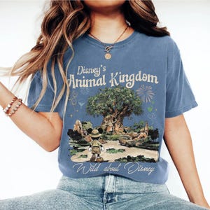 May include: Blue t-shirt featuring Disney's Animal Kingdom. The design includes the Animal Kingdom logo, a tree, Mickey Mouse in safari attire, and the phrase "Wild about Disney". The shirt is a comfortable, casual wear.
