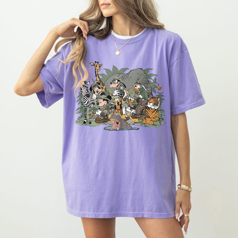 K&ouml;nnte beinhalten: Ein lilafarbenes T-Shirt mit einer farbenfrohen Cartoon-Illustration verschiedener Safari-Tiere und Disney-Figuren. Das Design umfasst ein Zebra, eine Giraffe, einen Elefanten, einen Tiger und Micky Maus, vor einem Hintergrund aus gr&uuml;nem Laub.