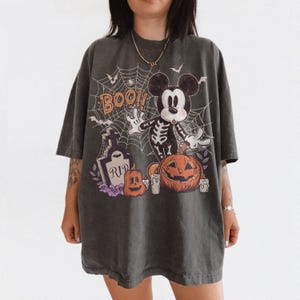 May include: A charcoal grey oversized t-shirt featuring a graphic print of a skeleton-costumed mouse character, a tombstone with the text RIP, orange pumpkins, white candles, and a spiderweb background with the orange text BOO.