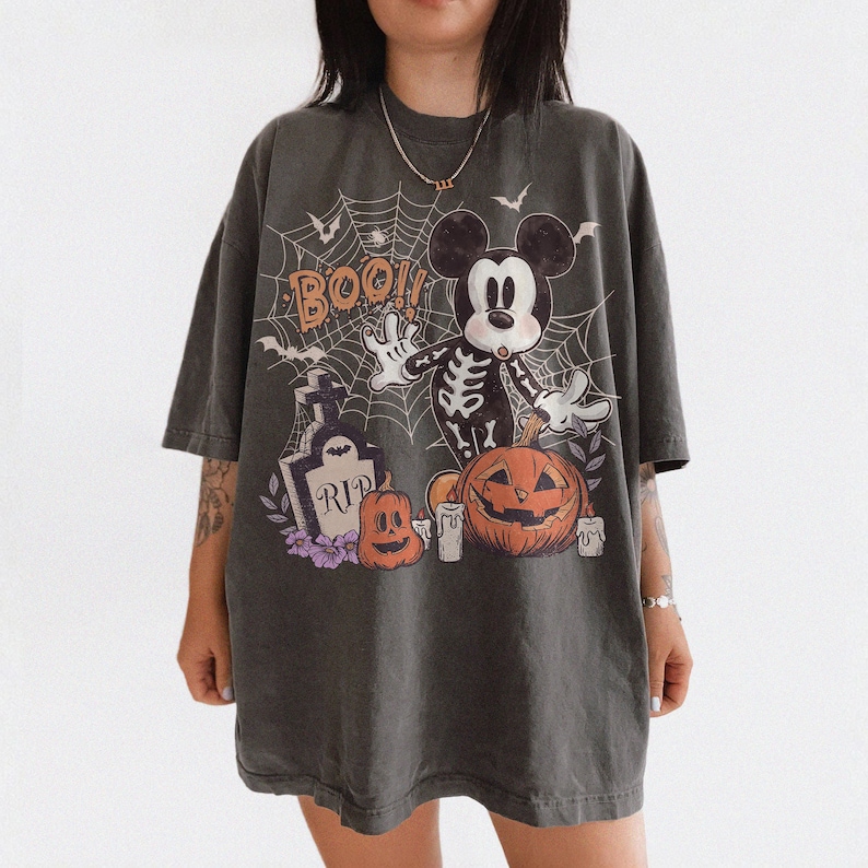 May include: A charcoal grey oversized t-shirt featuring a graphic print of a skeleton-costumed mouse character, a tombstone with the text RIP, orange pumpkins, white candles, and a spiderweb background with the orange text BOO.