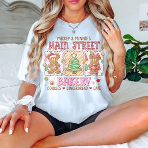 Op de afbeelding: Wit T-shirt met een Main Street Bakery-ontwerp met Mickey en Minnie Mouse peperkoekpersonages en een kerstboom. De tekst bevat "Mickey & Minnie's Main Street Bakery" en "Cookies, Gingerbread, Cake."