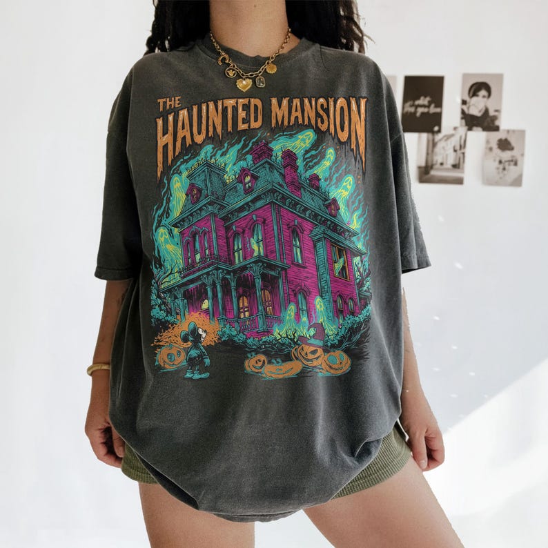 May include: Dark gray t-shirt featuring a colorful graphic of "The Haunted Mansion." The design includes a pink house, green and blue flames, and jack-o'-lanterns. The shirt has short sleeves and a relaxed fit, perfect for Halloween.