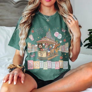 K&ouml;nnte beinhalten: Gr&uuml;nes T-Shirt mit einem Disneyland Main Street Trolley-Design. Das Design zeigt Micky Maus, Minnie Maus, Donald Duck und Goofy, sowie Feuerwerk, ein Schloss und das Wort "Disneyland".