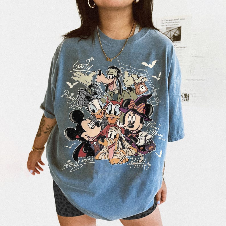 Może przedstawiać: Niebieski t-shirt z kr&oacute;tkim rękawem z nadrukiem graficznym przedstawiającym postacie Disneya w kostiumach na Halloween, w tym Myszkę Miki, Myszkę Minnie, Goofy'ego, Daisy, Donalda i Pluto, z pajęczynami, nietoperzami i imionami postaci.