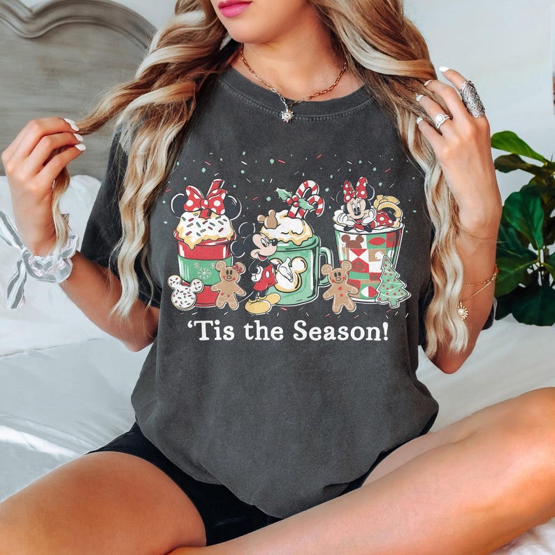 Puede incluir: Camiseta gris oscuro con un gr&aacute;fico festivo con los personajes de Mickey y Minnie Mouse, hombres de jengibre y bebidas tem&aacute;ticas navide&ntilde;as. La frase "'Tis the Season!" est&aacute; impresa debajo. La camiseta es de estilo casual.