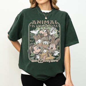 May include: Dark green t-shirt featuring a graphic of Disney characters, including Mickey and Minnie Mouse, Donald and Daisy Duck, and Goofy. The graphic is framed by the words "ANIMAL KINGDOM" and "Walt Disney World". The shirt has a vintage, worn look.