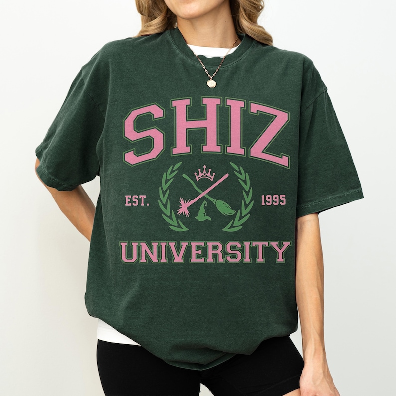 Pu&ograve; includere: T-shirt verde scuro con la scritta "SHIZ UNIVERSITY" in rosa. La maglietta presenta uno stemma con un cappello da strega, bacchette incrociate e l'anno "1995". La maglietta ha un girocollo e maniche corte.