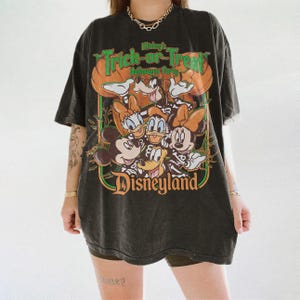 K&ouml;nnte beinhalten: Ein schwarzes Kurzarm-T-Shirt mit einem Grafikdruck von Disney-Figuren in Skelettkost&uuml;men. Der Text lautet Mickey's Trick-or-Treat Halloween Party und Disneyland. Das Design enth&auml;lt orangefarbene K&uuml;rbisse und gr&uuml;ne Akzente auf dunklem Grund.