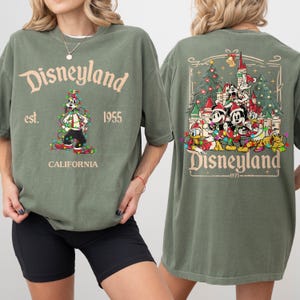 May include: Olive green Disneyland t-shirt. The front features the text "Disneyland est. 1955 CALIFORNIA" with a graphic of Goofy in Christmas lights. The back has a Disneyland graphic with Mickey, Minnie, Donald, and Goofy in Santa hats.