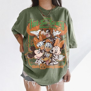 May include: An olive green t-shirt featuring a graphic print of Mickey Mouse, Minnie Mouse, Donald Duck, Daisy Duck, and Goofy wearing skeleton costumes. The text reads Mickey's Not-So-Scary Halloween Party and Walt Disney World in orange and green.