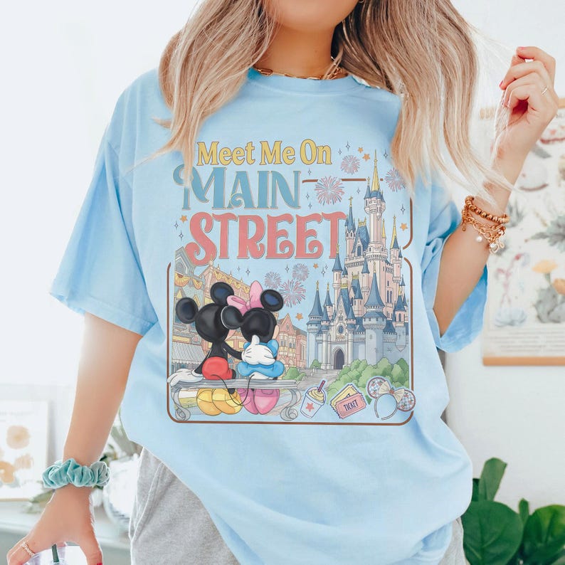 Op de afbeelding: Lichtblauw T-shirt met een afbeelding van Mickey en Minnie Mouse op een bankje voor een kasteel. De tekst "Meet Me On Main Street" staat in een retro lettertype boven de afbeelding. Vuurwerk en andere details maken het ontwerp compleet.