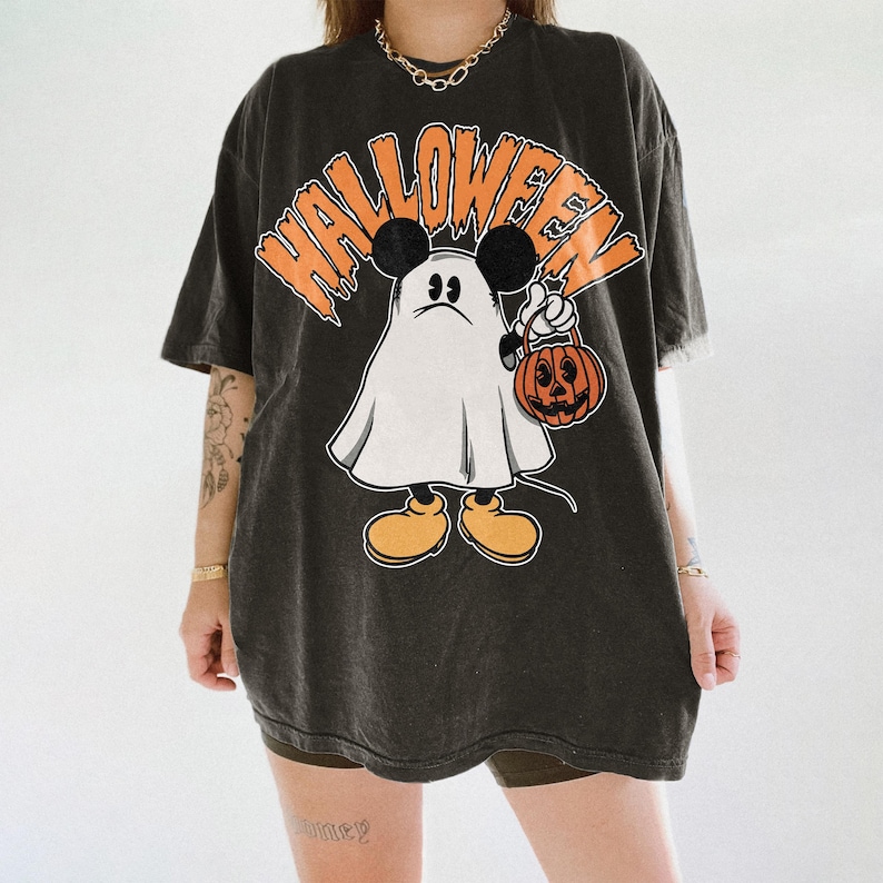 May include: Dark gray t-shirt with a Halloween-themed graphic. The design features a cartoon ghost character with Mickey Mouse ears holding a jack-o'-lantern. The word "HALLOWEEN" is printed above the character in orange.