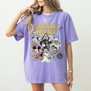 May include: A lilac-colored t-shirt featuring the "Star Wars" logo and cartoon characters such as Mickey Mouse, Minnie Mouse, Donald Duck, and Goofy dressed as Star Wars characters. The shirt has a relaxed fit.