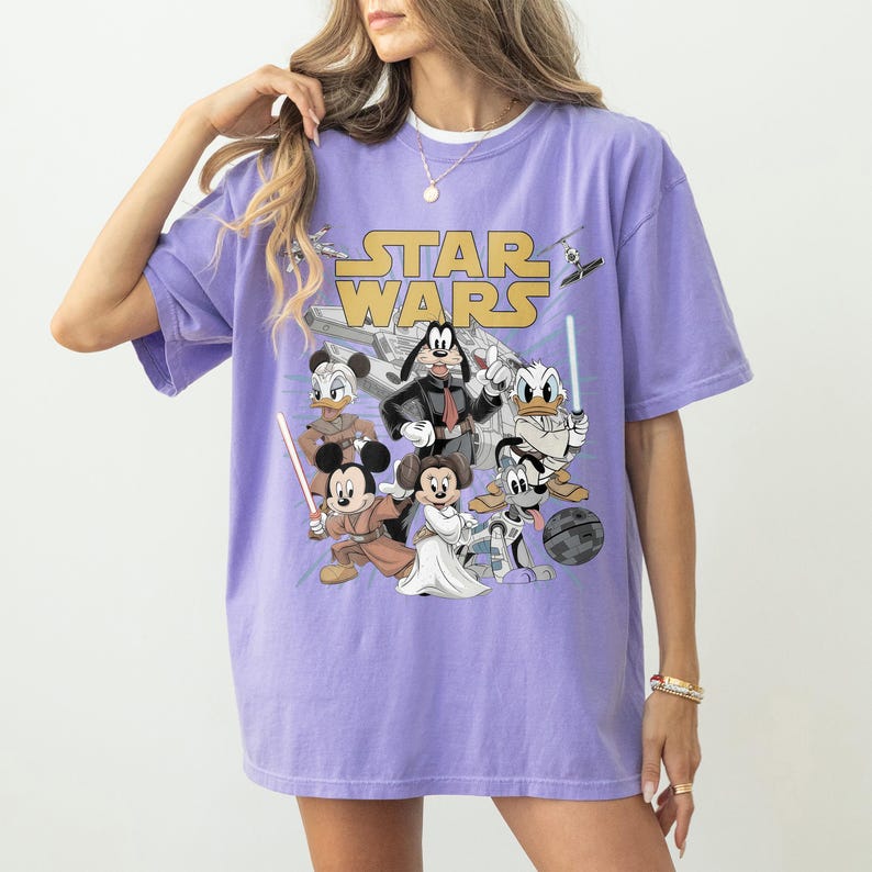 May include: A lilac-colored t-shirt featuring the "Star Wars" logo and cartoon characters such as Mickey Mouse, Minnie Mouse, Donald Duck, and Goofy dressed as Star Wars characters. The shirt has a relaxed fit.