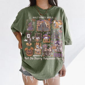 May include: Olive green t-shirt featuring a graphic design for Mickey's Not-So-Scary Halloween Party at Walt Disney World. The design includes cartoon characters, a castle, and the text "2020".