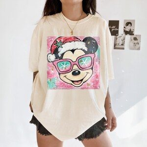K&ouml;nnte beinhalten: Cremefarbenes T-Shirt mit einer Grafik von Mickey Mouse mit Weihnachtsm&uuml;tze und rosa Sonnenbrille. Das Design hat einen rosa-gr&uuml;nen Aquarellhintergrund mit Sternakzenten. Eine goldene Halskette ist sichtbar.