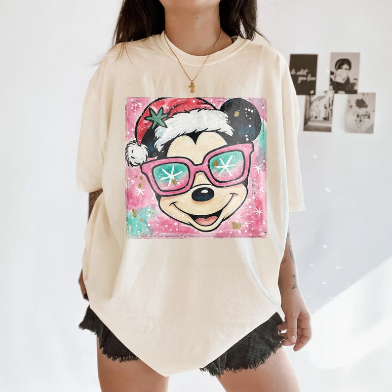 K&ouml;nnte beinhalten: Cremefarbenes T-Shirt mit einer Grafik von Mickey Mouse mit Weihnachtsm&uuml;tze und rosa Sonnenbrille. Das Design hat einen rosa-gr&uuml;nen Aquarellhintergrund mit Sternakzenten. Eine goldene Halskette ist sichtbar.