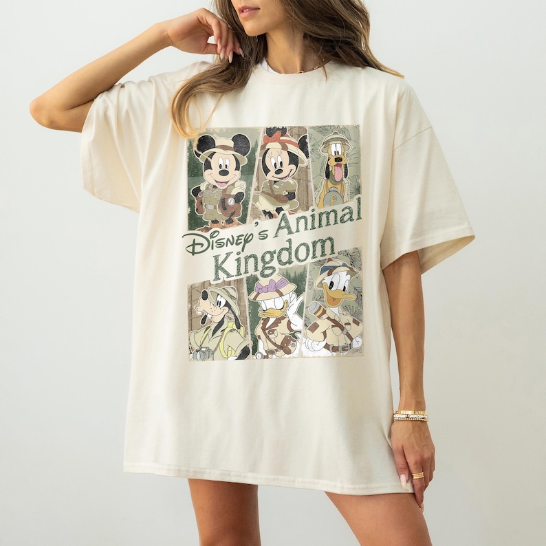 May include: Cream-colored t-shirt featuring a graphic of Disney's Animal Kingdom characters, including Mickey Mouse, Minnie Mouse, and Goofy. The text "Disney's Animal Kingdom" is printed in green. The shirt has short sleeves and a relaxed fit.