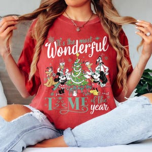 May include: Red t-shirt with a Christmas-themed graphic featuring Mickey Mouse, Minnie Mouse, Donald Duck, and Goofy around a decorated Christmas tree. The text on the shirt reads "It's the most Wonderful TIME of the year."