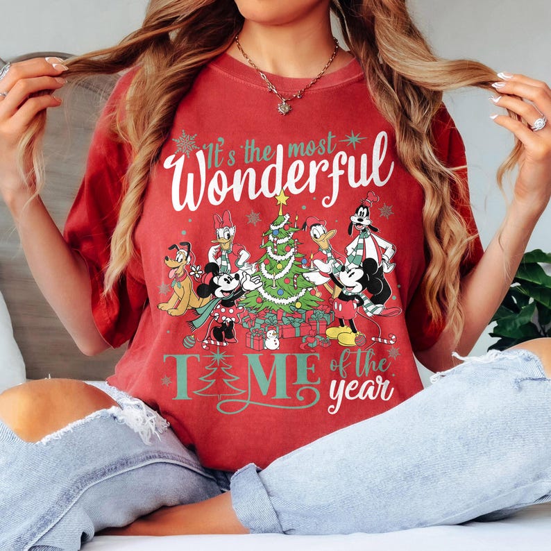 May include: Red t-shirt with a Christmas-themed graphic featuring Mickey Mouse, Minnie Mouse, Donald Duck, and Goofy around a decorated Christmas tree. The text on the shirt reads "It's the most Wonderful TIME of the year."