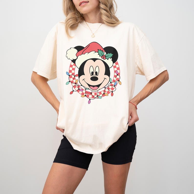 May include: Cream-colored t-shirt featuring a festive Mickey Mouse graphic. Mickey is wearing a Santa hat and surrounded by a wreath of Christmas lights. The design includes a red and white checkered pattern. The shirt has short sleeves.