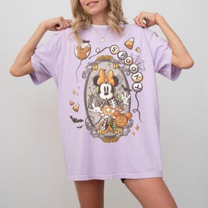May include: A light purple t-shirt featuring a Halloween-themed Minnie Mouse graphic. The design includes a skeleton Minnie, a pumpkin, candy corn, and the words "Spotlight". The shirt has short sleeves and a relaxed fit.