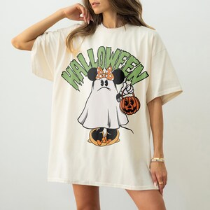 May include: Cream-colored t-shirt with a Halloween-themed graphic. The design features a cartoon character dressed as a ghost, holding a jack-o'-lantern. The word "HALLOWEEN" is printed above the character in green block letters.