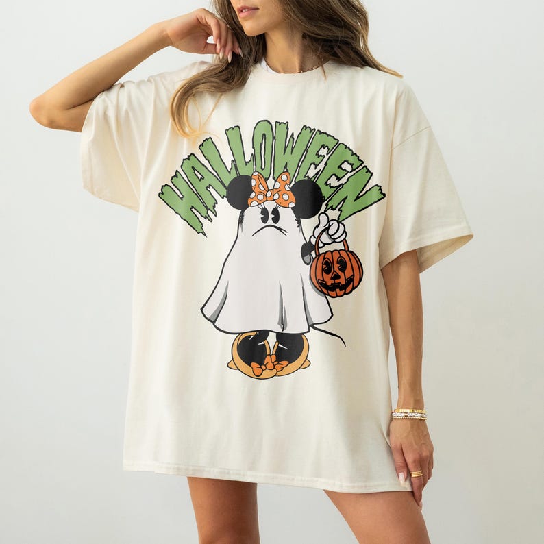 May include: Cream-colored t-shirt with a Halloween-themed graphic. The design features a cartoon character dressed as a ghost, holding a jack-o'-lantern. The word "HALLOWEEN" is printed above the character in green block letters.