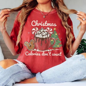 May include: Red Christmas-themed t-shirt with a graphic of a hot chocolate mug, gingerbread cookie, and Christmas tree. The text "Christmas Calories don't count" is printed on the shirt. The shirt is a short-sleeved crew neck.