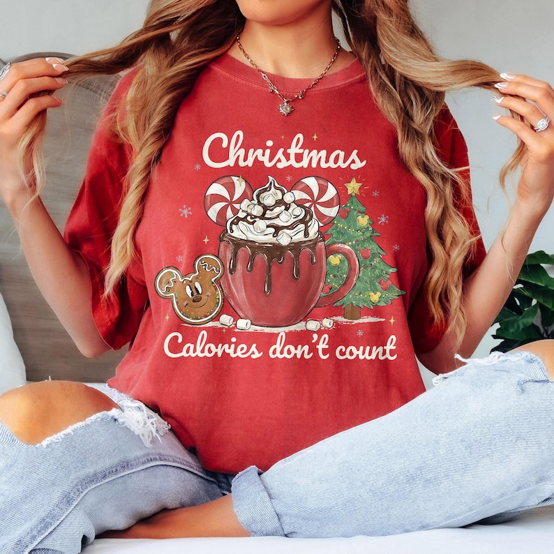 May include: Red Christmas-themed t-shirt with a graphic of a hot chocolate mug, gingerbread cookie, and Christmas tree. The text "Christmas Calories don't count" is printed on the shirt. The shirt is a short-sleeved crew neck.
