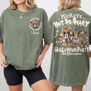 May include: Olive green t-shirt featuring a Halloween-themed graphic. The front displays a skeleton Goofy with a pumpkin and the word "Uncle." The back showcases Mickey Mouse and friends as skeletons, with the text "Mickey's Not So Scary Halloween Party" and "Walt Disney World."