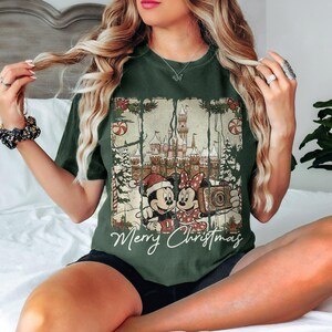 May include: Dark green t-shirt featuring a Christmas-themed graphic with Mickey and Minnie Mouse, a castle, and the words "Merry Christmas." The design includes candy canes and Christmas trees. The shirt is a comfortable, casual style.