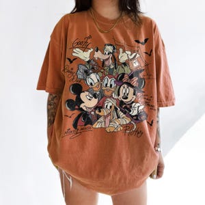 Może przedstawiać: T-shirt z kr&oacute;tkim rękawem w kolorze rdzawym z nadrukiem graficznym przedstawiającym postacie Disneya w kostiumach na Halloween, w tym Myszka Miki, Myszka Minnie, Goofy, Kaczor Donald, Kaczka Daisy i Pluto, z napisami przy każdej postaci.
