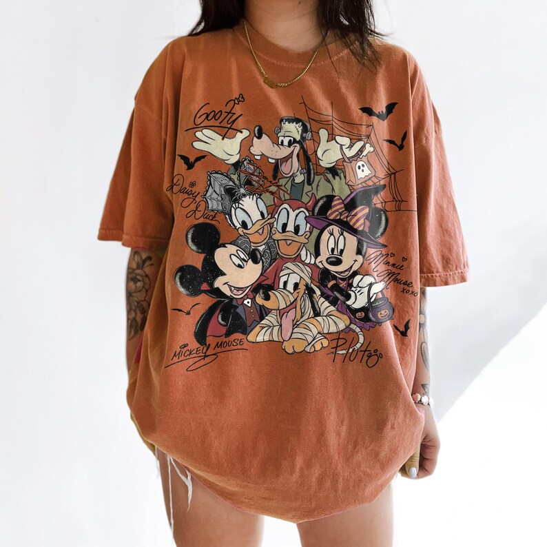 Może przedstawiać: T-shirt z kr&oacute;tkim rękawem w kolorze rdzawym z nadrukiem graficznym przedstawiającym postacie Disneya w kostiumach na Halloween, w tym Myszka Miki, Myszka Minnie, Goofy, Kaczor Donald, Kaczka Daisy i Pluto, z napisami przy każdej postaci.