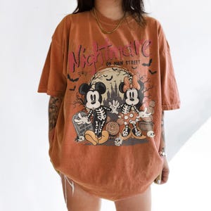 May include: An orange t-shirt with a graphic featuring Mickey and Minnie Mouse dressed as skeletons in front of a spooky scene. The text "Nightmare on Main Street" is printed across the top in pink and black.