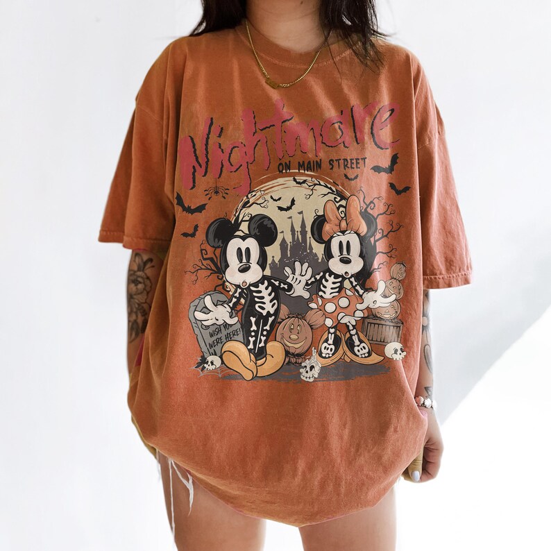 May include: An orange t-shirt with a graphic featuring Mickey and Minnie Mouse dressed as skeletons in front of a spooky scene. The text "Nightmare on Main Street" is printed across the top in pink and black.