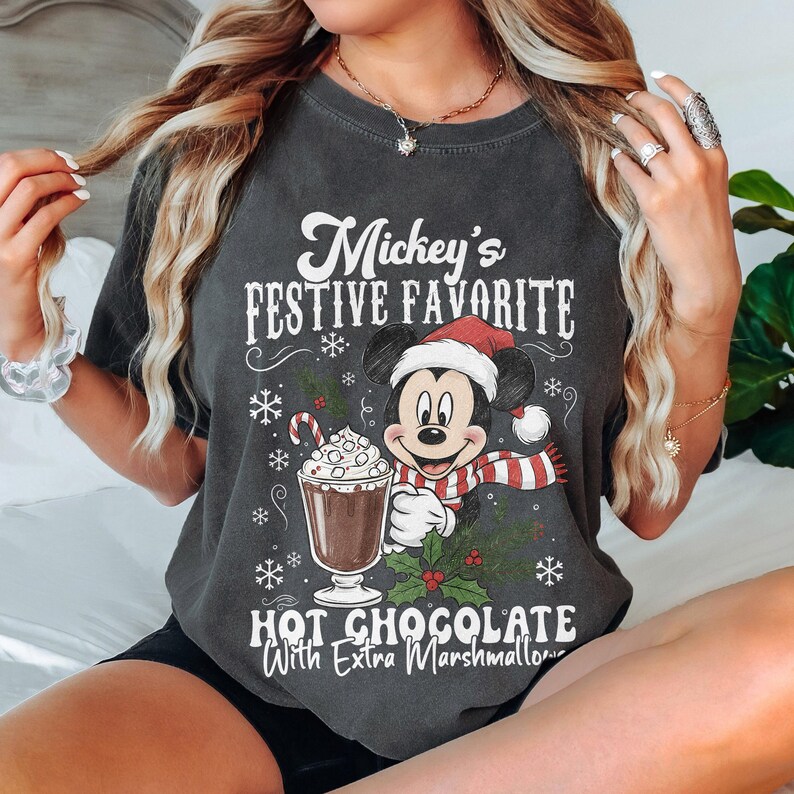 May include: Dark gray t-shirt featuring Mickey Mouse in a Santa hat and scarf, holding a cup of hot chocolate with marshmallows. The text reads "Mickey's Festive Favorite Hot Chocolate With Extra Marshmallows."