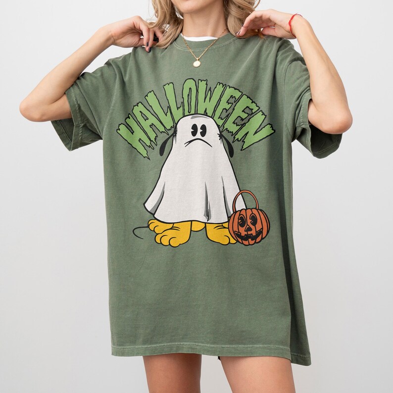 May include: Olive green Halloween-themed t-shirt featuring a cartoon ghost character with a sad expression. The word "HALLOWEEN" is printed above the ghost in green, stylized lettering. The ghost is holding a jack-o'-lantern bucket. The shirt has short sleeves.