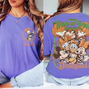 May include: A purple t-shirt featuring a front graphic of Daisy Duck in a skeleton costume inside a checkered circle with the word Auntie. The back displays Mickey, Minnie, Donald, Daisy, and Goofy in skeleton costumes with the text Mickey's Trick-or-Treat Halloween Party Disneyland.