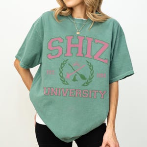 Pu&ograve; includere: Una maglietta verde con la scritta "SHIZ UNIVERSITY" in rosa. Il design include uno stemma con bacchette incrociate e una corona d'alloro. La maglietta ha maniche corte e girocollo.