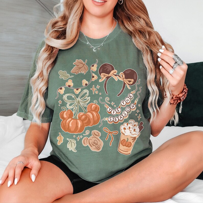 May include: An olive green short-sleeve t-shirt featuring a graphic print of orange pumpkins, brown mouse ears with bows, a beaded necklace with the text 'TIS THE SEASON, and a coffee cup with whipped cream and a pumpkin design in a fall theme.