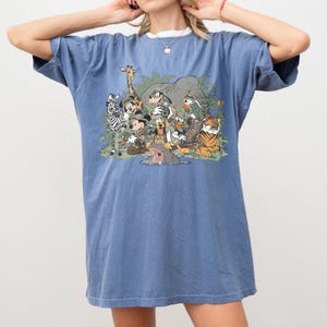 K&ouml;nnte beinhalten: Blaues T-Shirt mit einem Aufdruck von Disney-Figuren, darunter Micky Maus, Donald Duck und Goofy, sowie einem Zebra, einer Giraffe und einem Tiger. Das Shirt hat kurze &Auml;rmel und einen Rundhalsausschnitt.