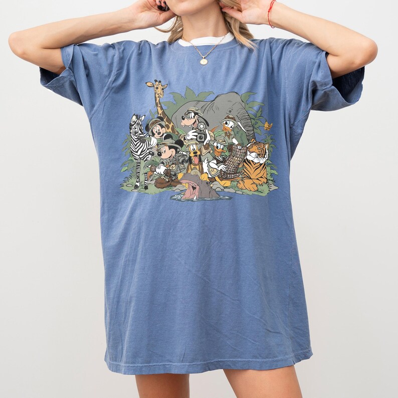 K&ouml;nnte beinhalten: Blaues T-Shirt mit einem Aufdruck von Disney-Figuren, darunter Micky Maus, Donald Duck und Goofy, sowie einem Zebra, einer Giraffe und einem Tiger. Das Shirt hat kurze &Auml;rmel und einen Rundhalsausschnitt.