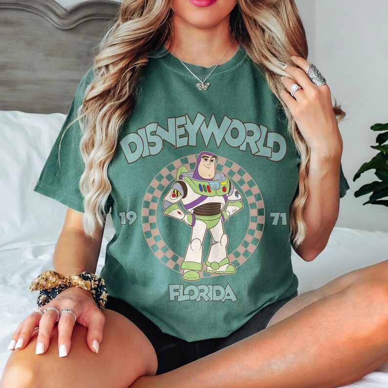 May include: Green t-shirt featuring a graphic of Buzz Lightyear from Disney's Toy Story, with the text "Disneyworld 1971 Florida". The design is set within a circular checkered pattern. The shirt is a solid green color.