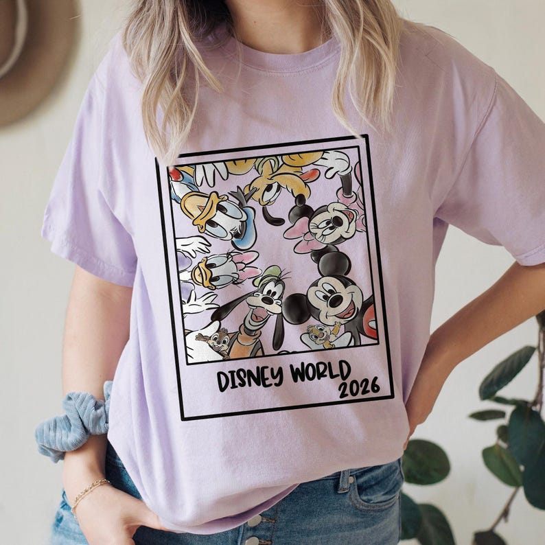 Puede incluir: Una camiseta lila con un dise&ntilde;o enmarcado de personajes de Disney, incluyendo Mickey y Minnie Mouse, Donald Duck y Goofy. El dise&ntilde;o lleva la inscripci&oacute;n "DISNEY WORLD 2026".