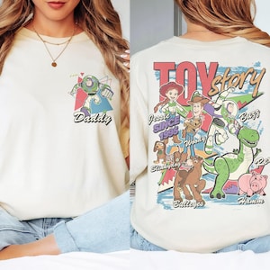 K&ouml;nnte beinhalten: Cremefarbenes T-Shirt mit Buzz Lightyear-Grafik und dem Wort "Daddy". Auf der R&uuml;ckseite befindet sich eine "Toy Story"-Grafik mit Figuren wie Woody, Jessie und Rex. Ein l&auml;ssiges Unisex-T-Shirt.