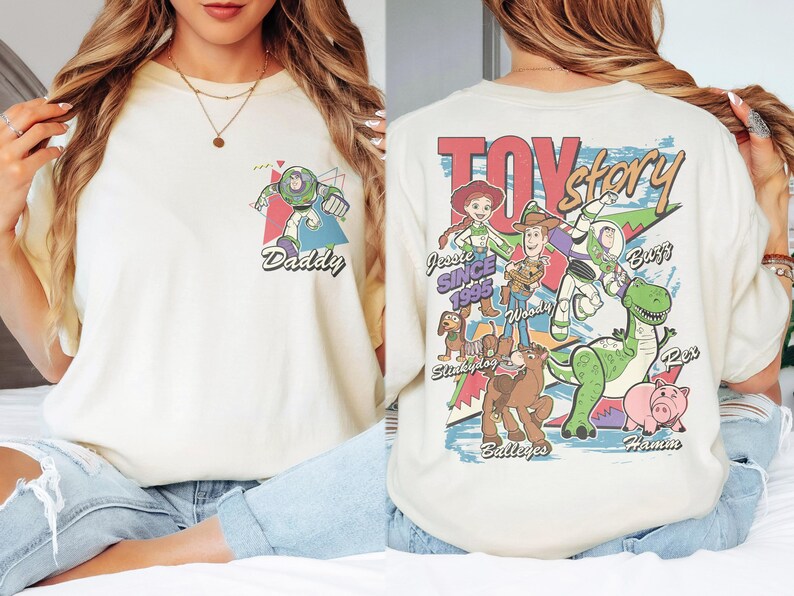 K&ouml;nnte beinhalten: Cremefarbenes T-Shirt mit Buzz Lightyear-Grafik und dem Wort "Daddy". Auf der R&uuml;ckseite befindet sich eine "Toy Story"-Grafik mit Figuren wie Woody, Jessie und Rex. Ein l&auml;ssiges Unisex-T-Shirt.